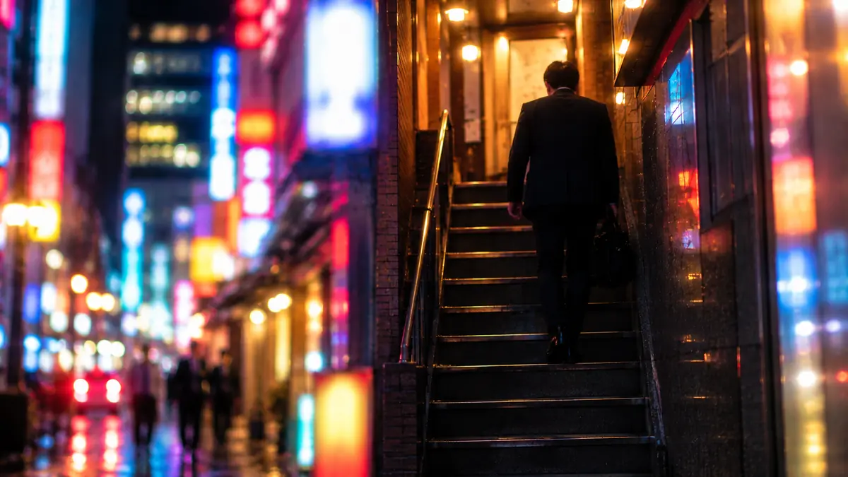 夜、繁華街の雑居ビルの入口の階段を上っていく人の後ろ姿